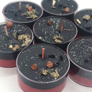 May include: A collection of black and red tea light candles. Each candle is decorated with small stones, herbs, and a small, unlit wick. The candles are arranged on a white surface.