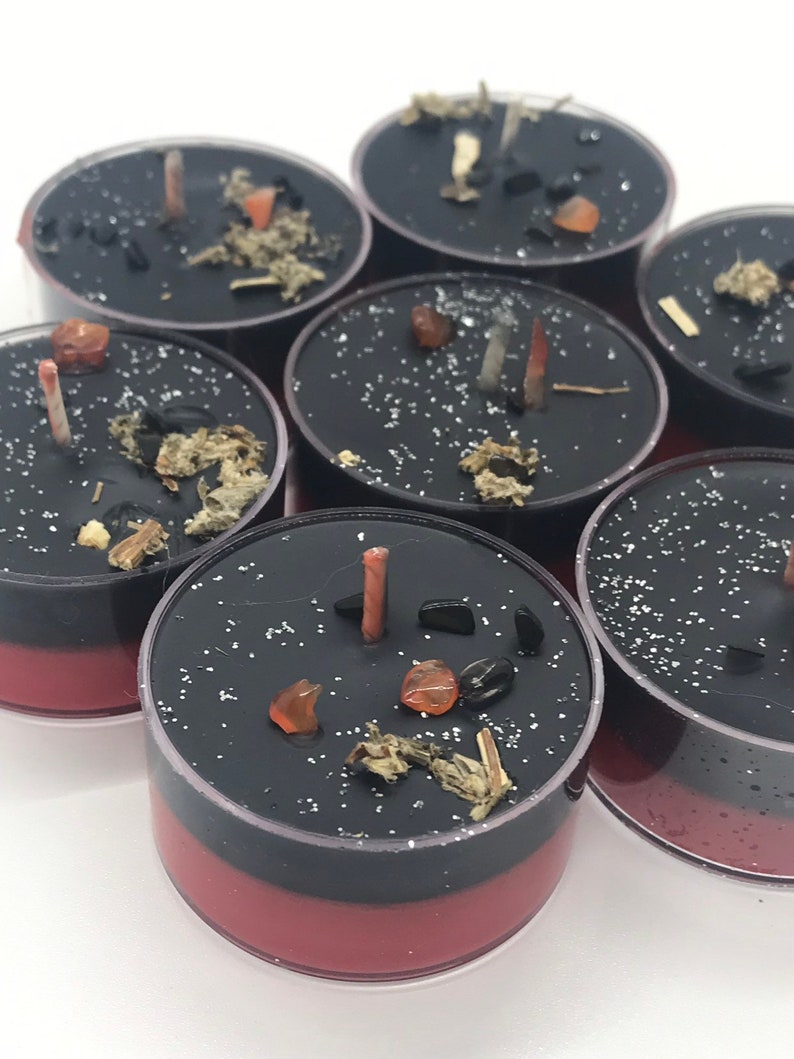 May include: A collection of black and red tea light candles. Each candle is decorated with small stones, herbs, and a small, unlit wick. The candles are arranged on a white surface.