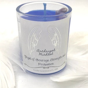 May include: A blue glass candle jar with a white label. The label features a pair of silver wings and the text "Archangel Michael Angel of Courage, Strength and Protection. Soy blend | Made in FL USA."