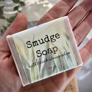 May include: A bar of soap wrapped in white paper with a floral design. The label reads "Smudge Soap with Black Tourmaline 3.2 oz".