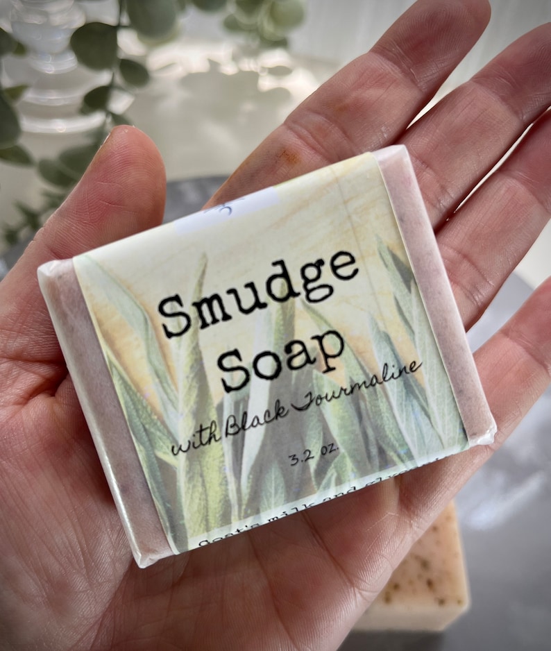 May include: A bar of soap wrapped in white paper with a floral design. The label reads "Smudge Soap with Black Tourmaline 3.2 oz".