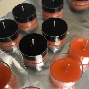 May include: A collection of tea light candles. Some candles are in clear plastic cups with orange wax. Other candles have black tops and orange wax. The candles are arranged on a reflective, silver surface.