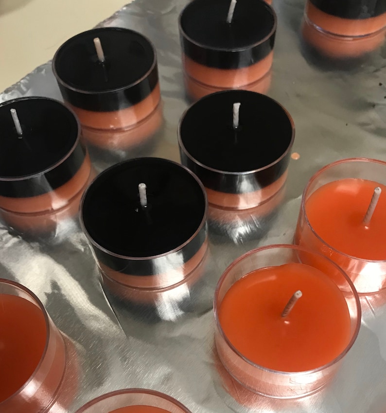 May include: A collection of tea light candles. Some candles are in clear plastic cups with orange wax. Other candles have black tops and orange wax. The candles are arranged on a reflective, silver surface.