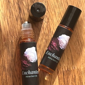 May include: Two small glass bottles with black roll-on tops and labels. The labels are black with white text that reads "Enchanted Attraction Oil" and feature a white rose with pink and purple accents.