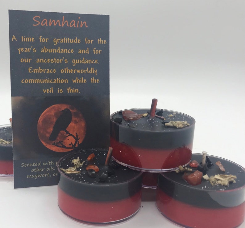 May include: Black and red tea light candles with decorative elements. A card with the word "Samhain" and text about gratitude and ancestor guidance is in the background. The candles are scented with mugwort and other oils.