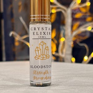 May include: A clear glass roll-on bottle with a gold cap, labeled "CRYSTAL ELIXIR 10ML." The label features a gold geometric crystal design and the words "BLOODSTON Strength & Grounding."