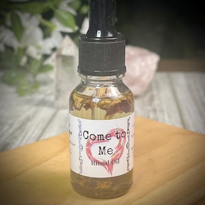 Puede incluir: Una pequeña botella de vidrio con un gotero en la parte superior llena de un líquido transparente y hierbas. La botella tiene una etiqueta blanca con el texto "Come to Me Ritual Oil" en un corazón rojo.