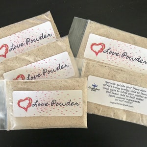 May include: Four small white bags with red hearts and the text "Love Powder" printed on them. The bags contain a beige powder. The text "Sprinkle outside your front door to attract loving energy. Add to laundry wash cycle to infuse your clothes and bed linens. Add to bath water. 100% natural ingredients. DO NOT INGEST. myenchantedpath.com" is printed on one of the bags.