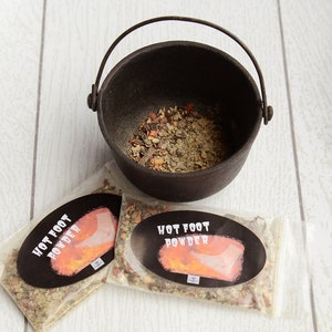May include: Two small clear plastic bags filled with a brown and green spice blend. The bags have black labels that read "Hot Foot Powder" with a graphic of a foot on fire. A small black cast iron pot with a handle is filled with the same spice blend.