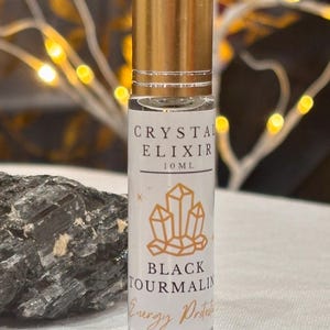 May include: A clear glass roller bottle with a gold cap, labeled "CRYSTAL ELIXIR 10 ML" and "BLACK TOURMALINE Energy Protection." The label features a gold crystal graphic. A dark gray stone sits beside the bottle, with a blurred background of fairy lights.
