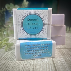 May include: Three bars of soap stacked on top of each other. The top bar has a blue and white label with the text "Cancel Clear Delete 3.2 ounce". The bottom bar has a blue and white label with the text "Those negative thoughts and emotions are not serving you! Take the time now to kick them to the curb."