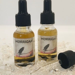 May include: Two small glass bottles with black dropper tops and labels that say "MORRIGAN" and "Magical Oil". A black crow silhouette is on each label. The bottles are sitting on a bed of clear quartz crystals.