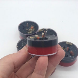 May include: A collection of black and red tea light candles. The candles have a two-tone design, with a black top and a red base. The candles are decorated with small pieces of herbs and crystals.