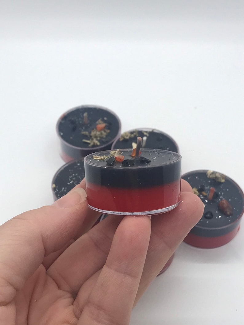 May include: A collection of black and red tea light candles. The candles have a two-tone design, with a black top and a red base. The candles are decorated with small pieces of herbs and crystals.