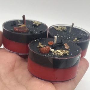 May include: Three small, layered candles in clear plastic cups. The bottom layer is red, topped with a black layer, and adorned with small crystals and herbs. Each candle has a wick. The candles are held in a hand.