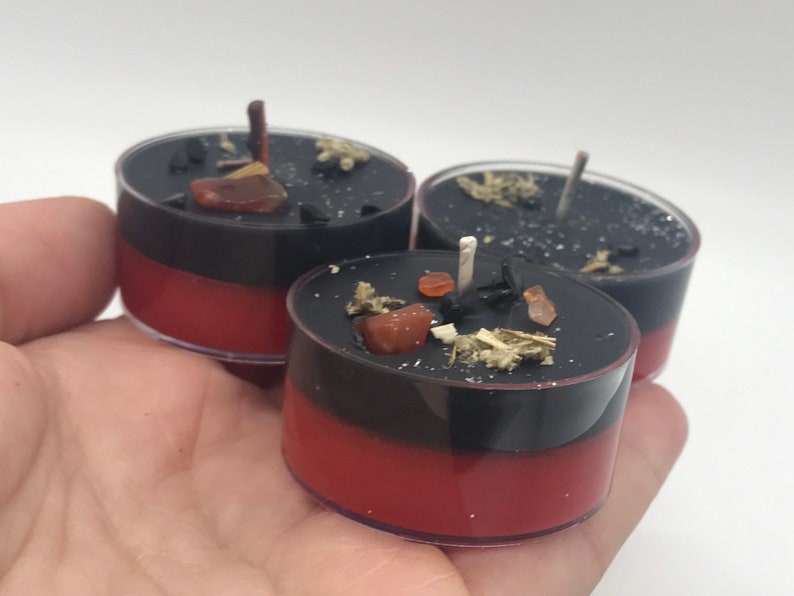 May include: Three small, layered candles in clear plastic cups. The bottom layer is red, topped with a black layer, and adorned with small crystals and herbs. Each candle has a wick. The candles are held in a hand.