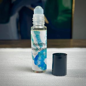 May include: A clear glass roller bottle with a black cap. The label is white with blue watercolor-style designs and the text "Healthy Boundaries" and "an oil for Empaths".
