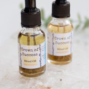 May include: Two small glass bottles with black droppers filled with a golden liquid. The bottles have white labels with the text "Crown of Success Ritual Oil".