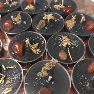 May include: A collection of black tea light candles, each adorned with a small gemstone, dried herbs, and a white wick. The candles are arranged in a circular pattern, creating a visually appealing display. The gemstones are a mix of orange and brown.