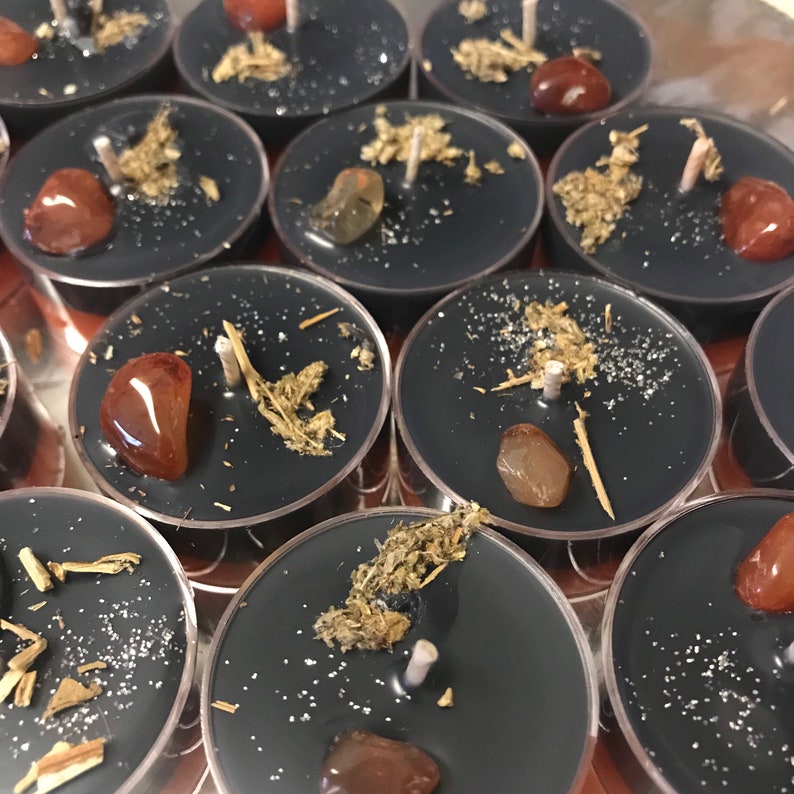 May include: A collection of black tea light candles, each adorned with a small gemstone, dried herbs, and a white wick. The candles are arranged in a circular pattern, creating a visually appealing display. The gemstones are a mix of orange and brown.