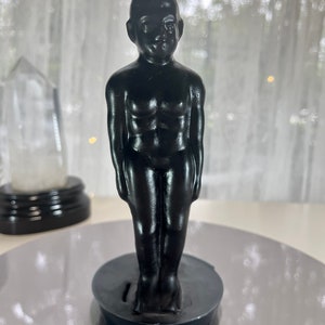 May include: A black candle shaped like a standing human figure. The candle has a detailed depiction of the torso and is mounted on a circular base. The candle is a solid black color and stands upright.