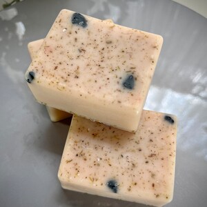 May include: Three bars of handmade soap with a light pink base and flecks of black and brown. The soap is stacked on top of each other.
