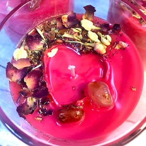 May include: A pink candle with a heart-shaped pool of melted wax. The candle is surrounded by dried rose petals, herbs, and crystals.