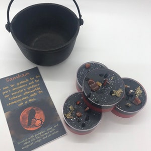 May include: A black cast iron cauldron with a handle sits next to four small, round candles. The candles have black wax, red rims, and are decorated with stones and herbs. A card with the word "Samhain" and text is also present.