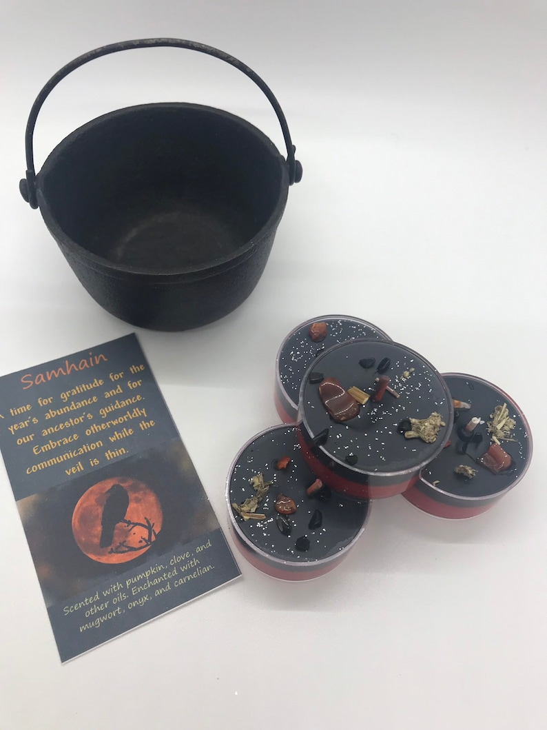 May include: A black cast iron cauldron with a handle sits next to four small, round candles. The candles have black wax, red rims, and are decorated with stones and herbs. A card with the word "Samhain" and text is also present.