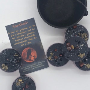 May include: A collection of black candles with embedded herbs and crystals, alongside a small cauldron and a card. The card reads "Samhain" and describes the theme of gratitude and guidance. The candles are scented with pumpkin, clove, and other oils.
