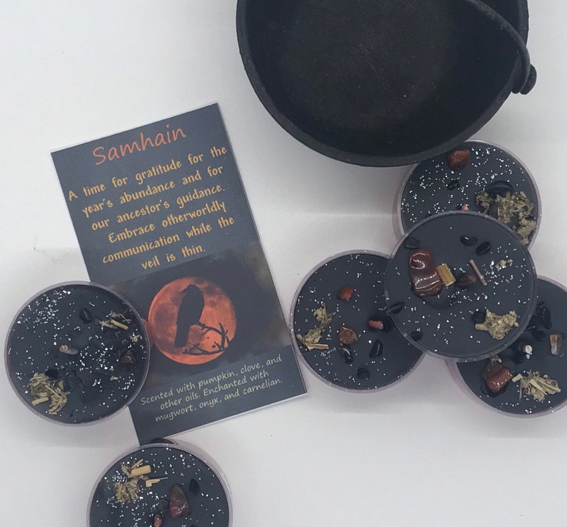May include: A collection of black candles with embedded herbs and crystals, alongside a small cauldron and a card. The card reads "Samhain" and describes the theme of gratitude and guidance. The candles are scented with pumpkin, clove, and other oils.