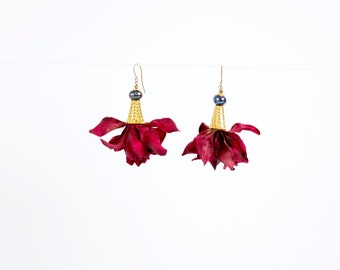 Puede incluir: Un par de pendientes con una flor de color rojo oscuro, una perla dorada y una perla negra en la parte superior.