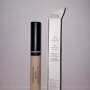 May include: A tube of bareMinerals Original Liquid Mineral Concealer with a black cap and a beige body. The product is next to its white box, which lists the product's features, including being vegan, gluten-free, and tree nut-free.