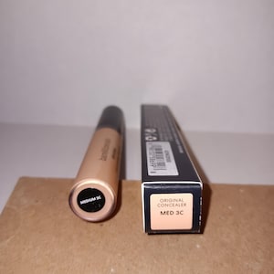 以下が含まれることがあります： ベージュ色のコンシーラーチューブとその箱。チューブの底には「MEDIUM 3C」と記載されています。箱には「ORIGINAL CONCEALER MED 3C」と書かれています。製品は茶色の表面にあります。