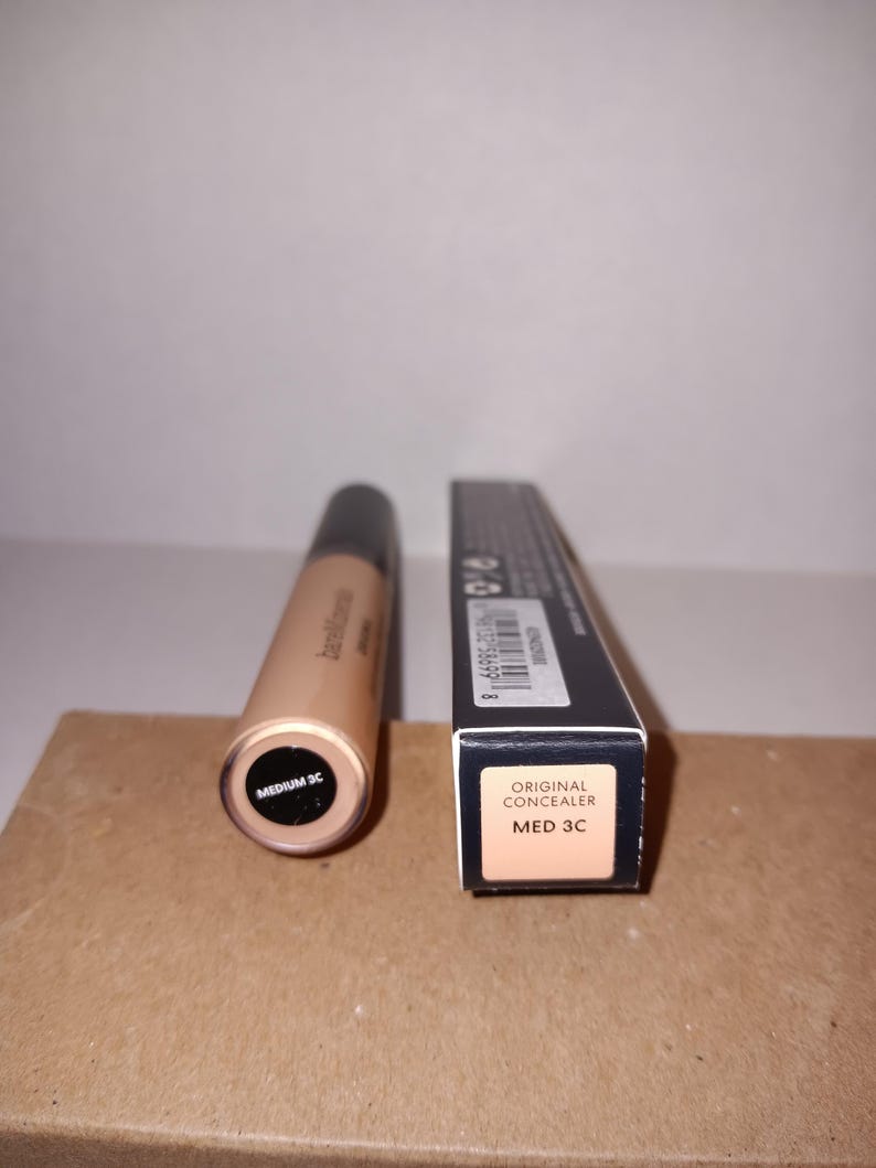 以下が含まれることがあります： ベージュ色のコンシーラーチューブとその箱。チューブの底には「MEDIUM 3C」と記載されています。箱には「ORIGINAL CONCEALER MED 3C」と書かれています。製品は茶色の表面にあります。