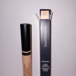 以下が含まれることがあります： ベージュ色のBareMinerals Original Liquid Mineral Concealerのチューブと、その黒い箱。箱には「Original Liquid Mineral Concealer」と「MED 3C」と書かれています。コンシーラーチューブは6 ml / 0.2 fl. oz.です。