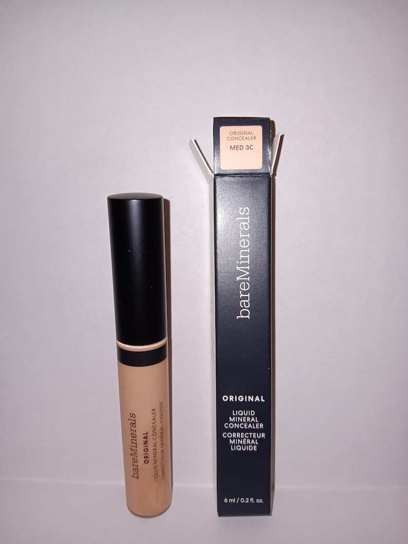 以下が含まれることがあります： ベージュ色のBareMinerals Original Liquid Mineral Concealerのチューブと、その黒い箱。箱には「Original Liquid Mineral Concealer」と「MED 3C」と書かれています。コンシーラーチューブは6 ml / 0.2 fl. oz.です。
