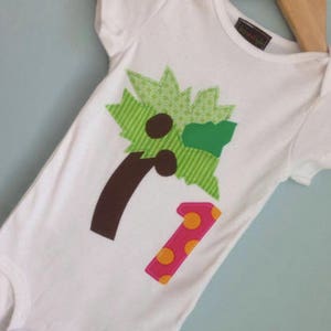 Pode incluir: Macacão branco para bebê com uma aplicação verde, marrom e rosa de uma palmeira e o número um.