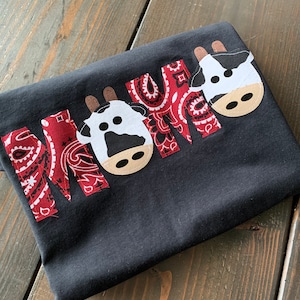 Może przedstawiać: Czarny T-shirt z napisem "MAMA" napisanym na czerwono-białym materiale bandany. Każda litera jest ozdobiona czarno-białą twarzą krowy.