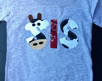 Puede incluir: Una camiseta gris de manga corta con la palabra "SIS" escrita en tela de estampado de vaca en blanco y negro. La "I" es un estampado de bandana rojo y negro.