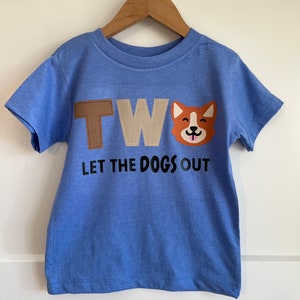 Op de afbeelding: Blauw T-shirt met korte mouwen met het woord "TWO" in bruine en beige viltletters en een cartoon-hondenkop. De tekst "LET THE DOGS OUT" is in zwart onder de viltletters gedrukt.