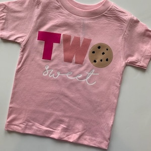 May include: Pink short-sleeved t-shirt with the text "TWO sweet" and a chocolate chip cookie graphic.