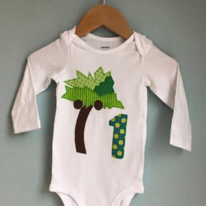 Pode incluir: Macacão branco de manga comprida com uma palmeira verde, marrom e amarela e o número um com bolinhas amarelas.