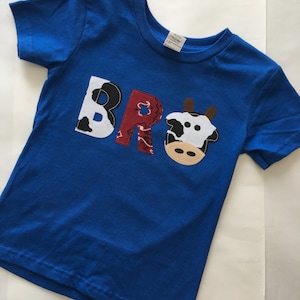 May include: A blue short-sleeved t-shirt with the word "BRO" spelled out in colorful fabric patches. The letters are made of black and white cow print, red bandana print, and a cow face.