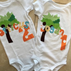 Pode incluir: Dois bodies brancos com aplicações verdes, marrons e laranja. Um body diz "Atticus 2" e o outro diz "Logan 1".