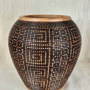May include: A handcrafted, decorative vase with a dark brown, geometric woven pattern. The vase has a light brown wooden rim and base. The design features a series of squares and rectangles, creating a textured surface. The vase is a unique home decor item.