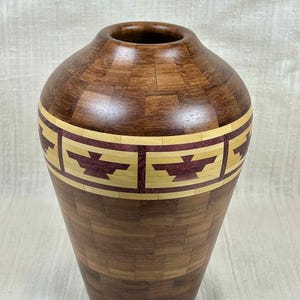 Può includere: Un vaso di legno fatto a mano con un corpo largo e un collo stretto. Il vaso è realizzato con varie tonalità di legno, con una fascia di disegni geometrici in crema e bordeaux. Il vaso è alto circa 25 cm.
