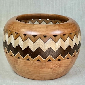 Può includere: Una ciotola in legno fatta a mano con un motivo a zig-zag unico. La ciotola presenta un mix di tonalità di legno chiare e scure, tra cui naturale, beige e marrone scuro. Il design della ciotola incorpora una serie di ritagli triangolari.