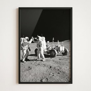 May include: Black and white framed print of two astronauts giving a high five on the moon. A lunar rover is parked nearby. The image captures a moment of celebration during a space mission.