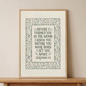 May include: A framed print featuring the text "BEFORE I FORMED YOU IN THE WOMB I KNEW YOU. BEFORE YOU WERE BORN I SET YOU APART. JEREMIAH 1:5." The text is in a decorative font, surrounded by a floral border with green, purple, and blue accents.